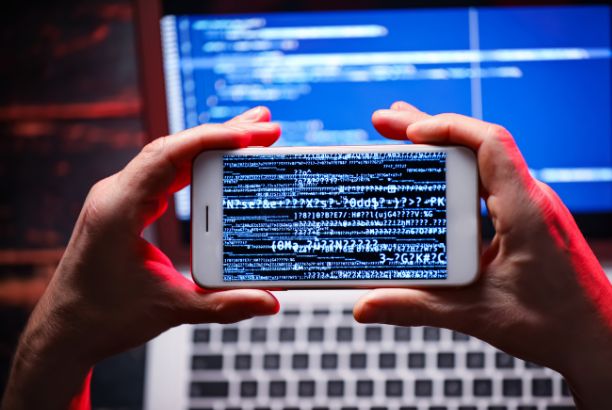 alt="Persona sosteniendo un móvil con código en la pantalla frente a un ordenador portátil con líneas de programación."