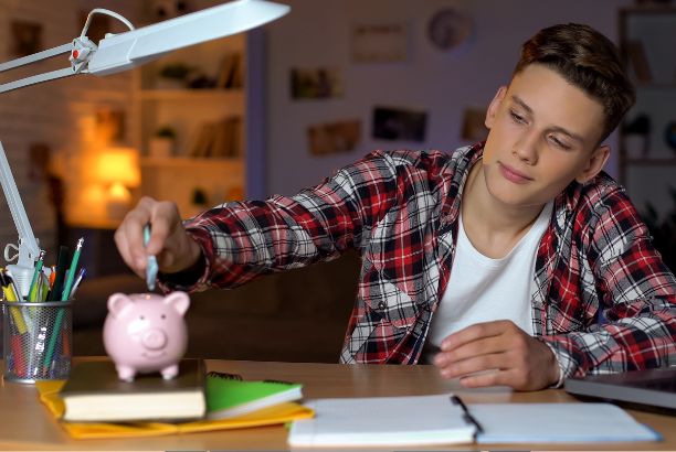 alt="Joven sentado en un escritorio colocando una moneda en una hucha en forma de cerdito, rodeado de libros y material de estudio.">