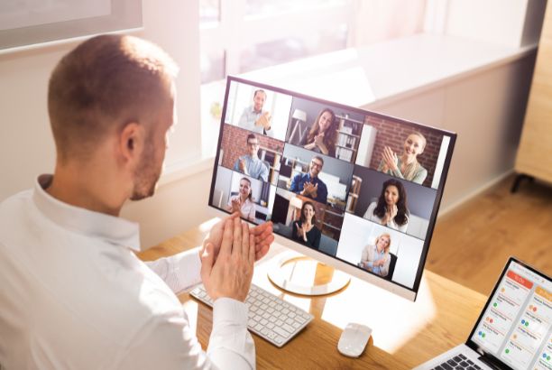 alt="Hombre participando en una videollamada grupal desde su ordenador de escritorio, con varias personas en pantalla aplaudiendo."