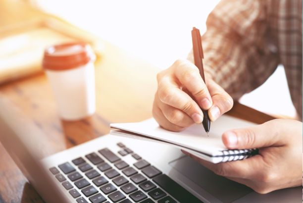 alt="Persona escribiendo en una libreta mientras usa un ordenador portátil, con una taza de café al fondo sobre la mesa."
